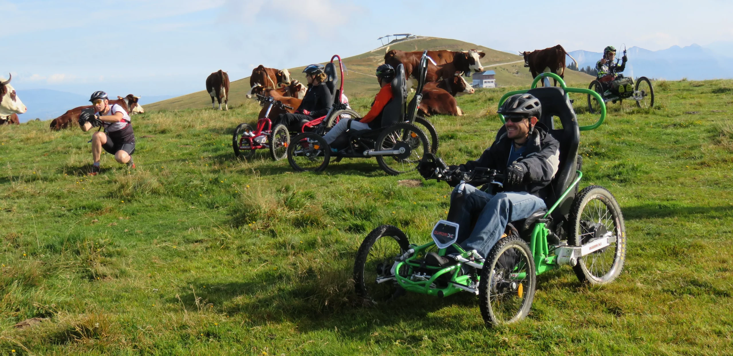 Quatre participants en fauteuil tout terrain et un accompagnateur au milieu des vaches sur le plateau du Semnoz pendant le R'Handi Bike Festival