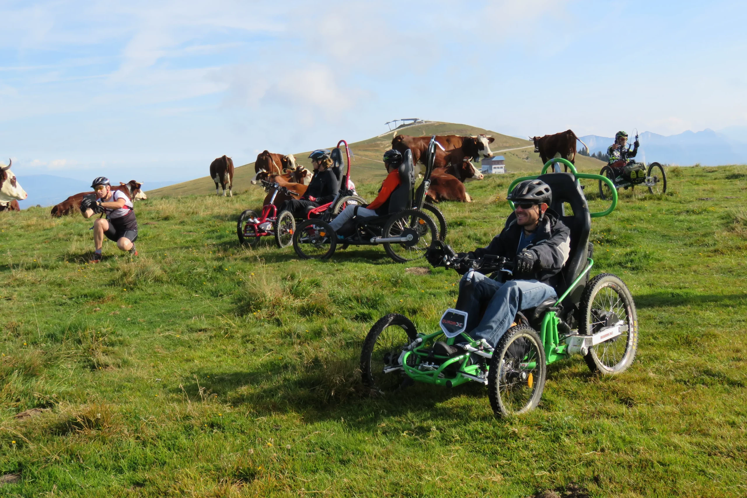 Quatre participants en fauteuil tout terrain et un accompagnateur au milieu des vaches sur le plateau du Semnoz pendant le R'Handi Bike Festival 2021