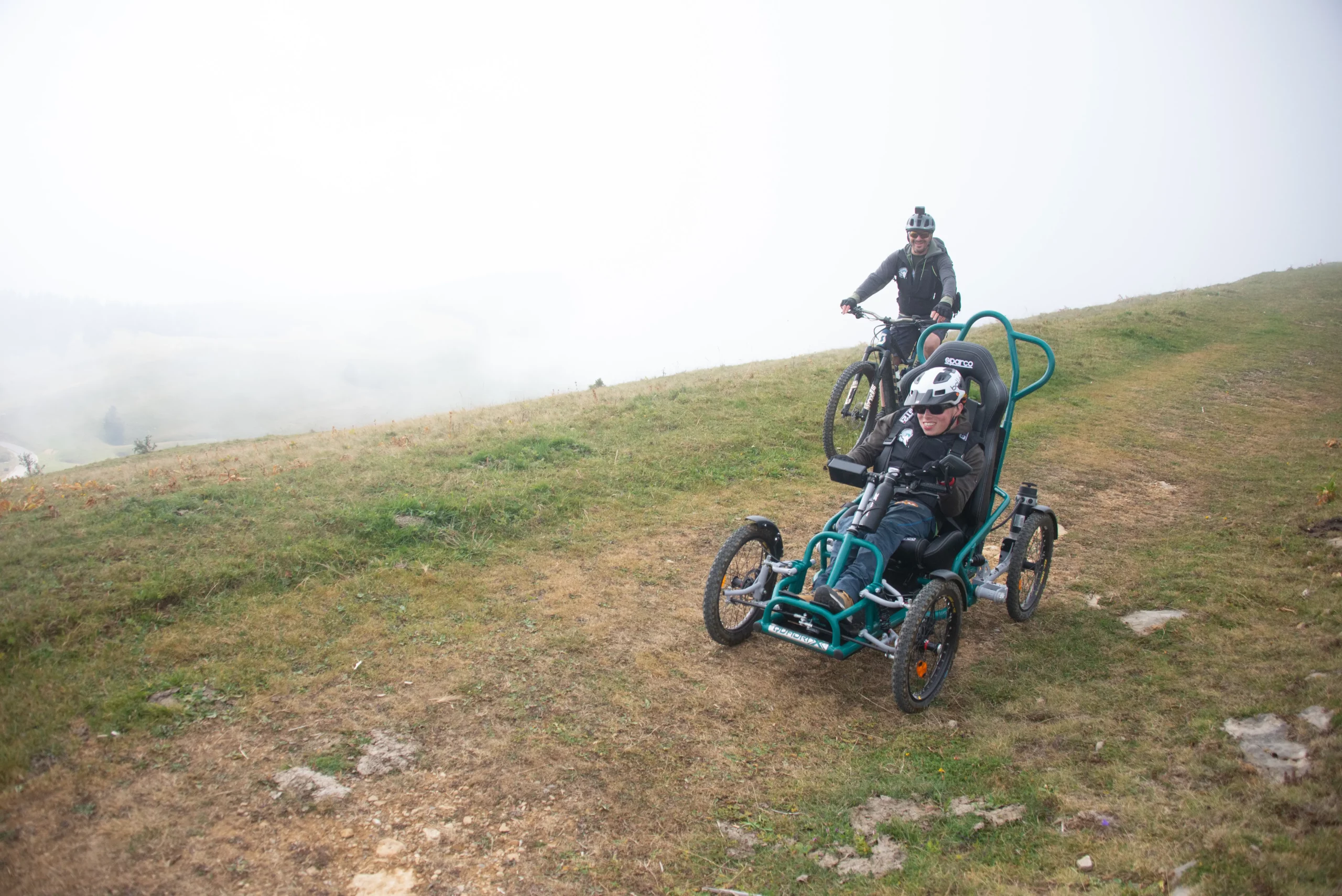 Gros plan d'un participant en FTT et son accompagnateur devant les nuages dans une descente sur chemin herbeux du Semnoz