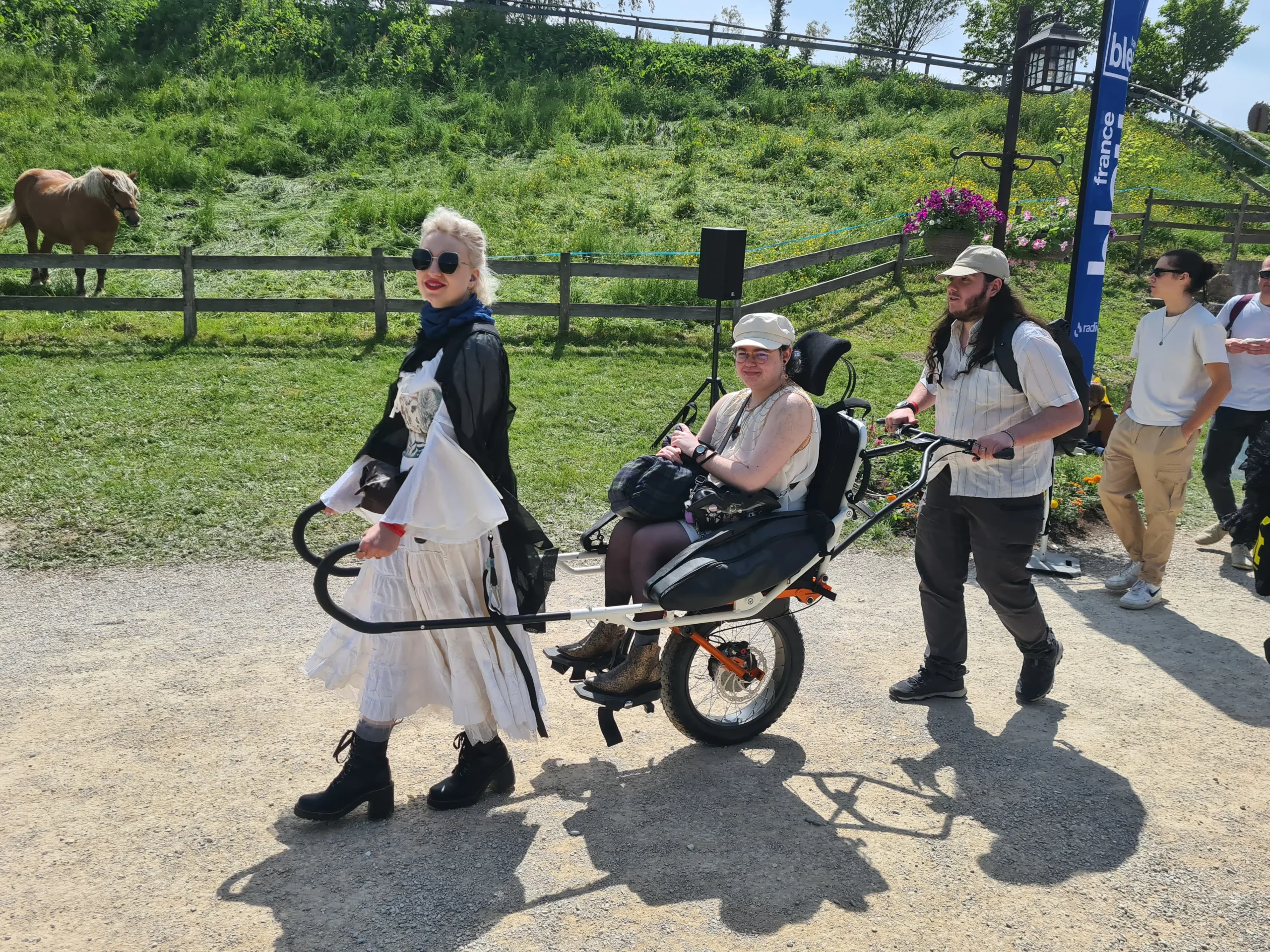 Deux accompagnateurs font découvrir les médiévales d'andilly à cette jeune femme grâce à une joëlette de l'association Handi Lac & Montagnes